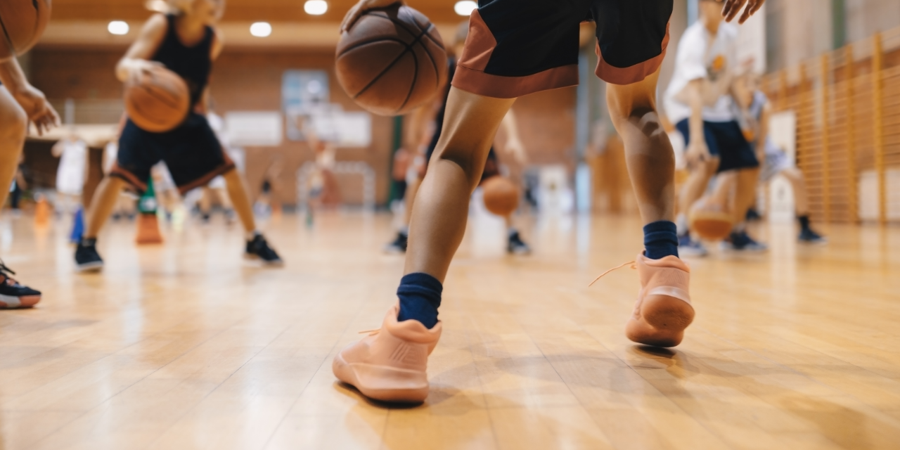 Basketball players dribbling on indoor court, showing legs and athletic shoes in motion.